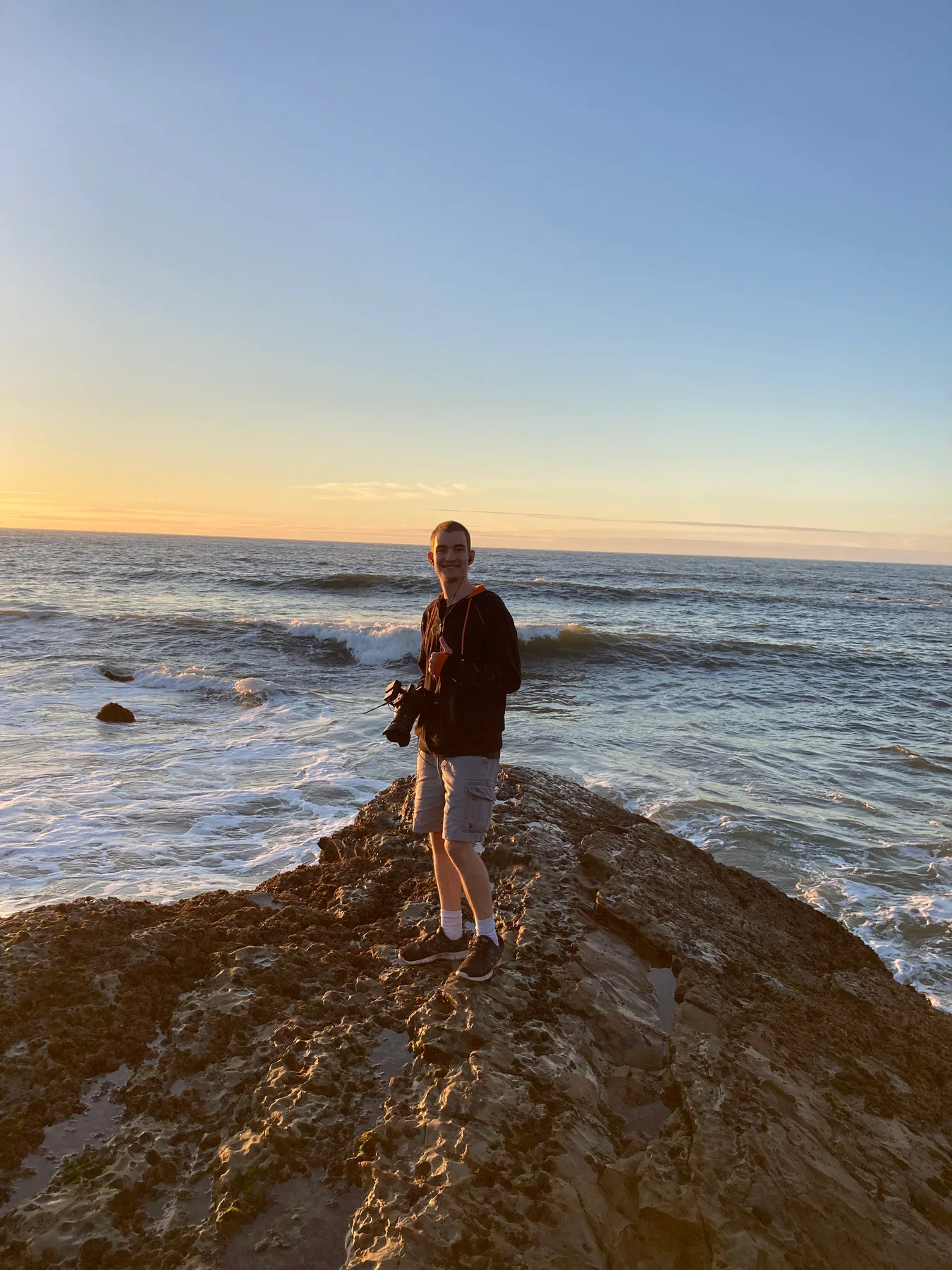 Zachary holding a camera on a rocky shoreline at sunset